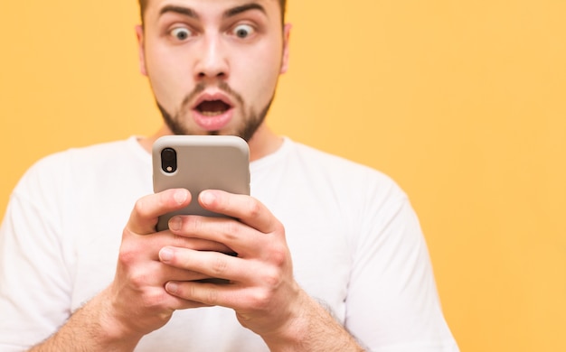 Erstaunter Teenager mit einem Bart, der ein Smartphone auf Gelb betrachtet