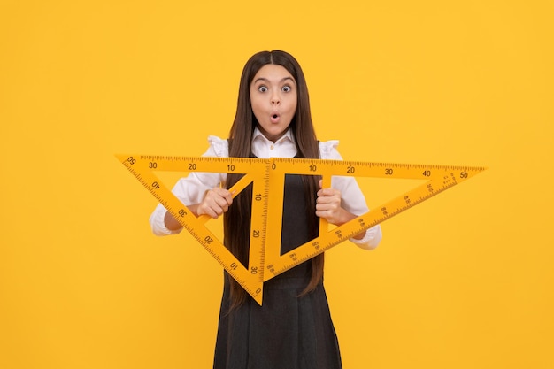 Erstauntes jugendlich Mädchen halten mathematisches Dreiecklineal in der Schule auf gelber Hintergrundbildung