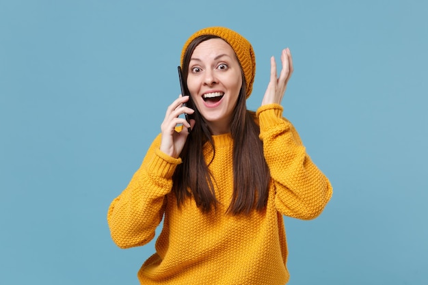 Erstauntes junges Brunettefrauenmädchen im gelben Pullover und im Hut, der lokalisiert auf blauem Hintergrundstudioporträt aufwirft. Menschen aufrichtige Emotionen Lifestyle-Konzept. Mock-up-Kopienbereich. Am Handy sprechen.