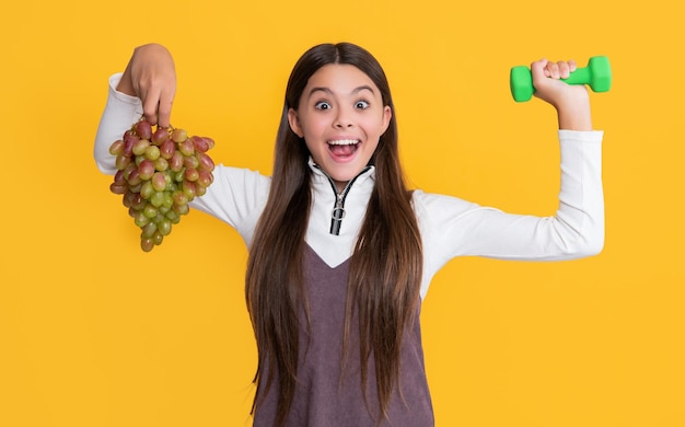 Erstauntes Kind hält frische Weintrauben und Sporthanteln auf gelbem Hintergrund