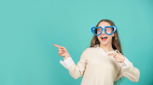 Erstauntes Kind in Partybrille, das mit dem Finger auf blauem Hintergrund mit Kopienraum zeigt, Modeaccessoire.