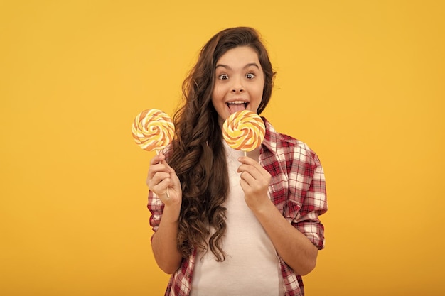 Erstauntes Kind mit langen lockigen Haaren im Hemd hält Lollipop-Karamellbonbons auf gelbem Hintergrund Kindheit