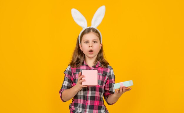 Erstauntes Osterkind in Hasenohren öffnet Geschenkbox auf gelbem Hintergrund