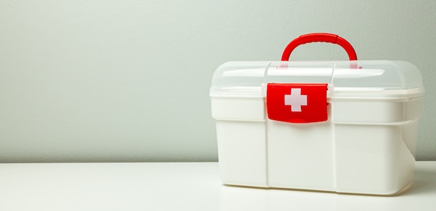 Erste-Hilfe-Kasten. Weiße Box mit Kreuz und rotem Verschluss auf grauem Grund.