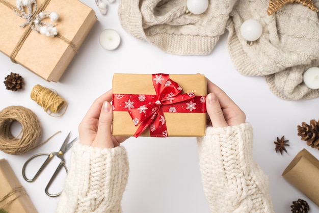 Erste-Person-Draufsichtfoto der Hände der Frau in einem kuscheligen Pullover, der eine Geschenkbox aus Bastelpapier mit roter Schleife über Schal-Weihnachtsbaumkugeln und Handwerkswerkzeugen auf isoliertem weißem Hintergrund hält