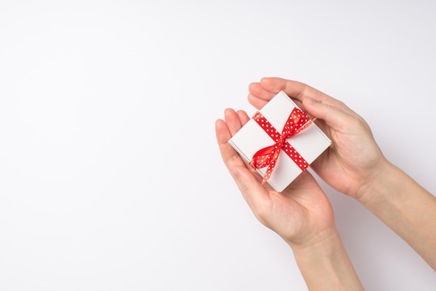 Erste-Person-Draufsichtfoto von Händen, die eine kleine weiße Geschenkbox mit rot gepunkteter Schleife auf isoliertem weißem Hintergrund mit leerem Raum geben