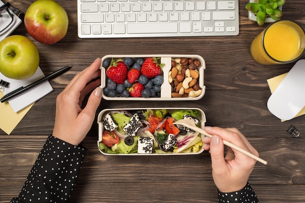 Erste-Person-Draufsichtfoto von Händen, die gesunde Lebensmittel essen, Salat, Beeren, Nüsse aus zwei Lunchboxen, Äpfeln, Glas mit Tastatur und Maus aus Saftpflanze, Schreibwaren auf isoliertem dunklem Holzschreibtischhintergrund