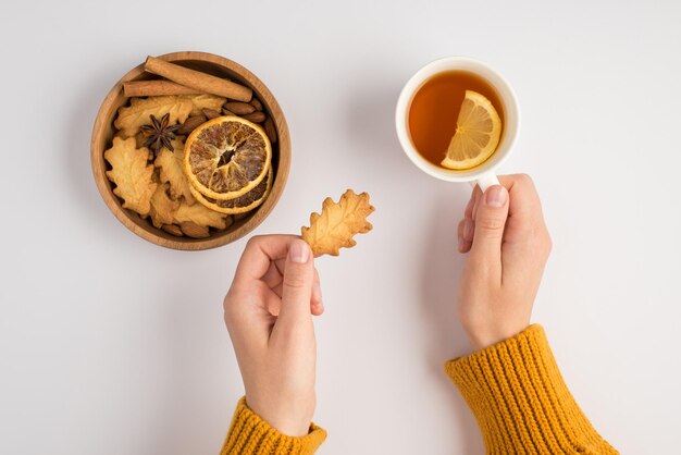 Erste-Person-Draufsichtfoto von weiblichen Händen im Pullover, die eine Tasse Tee und einen blattförmigen Keks über einer Holzschale mit Keksen, getrockneten Zitronenscheiben, Anis und Zimtstangen auf isoliertem weißem Hintergrund halten