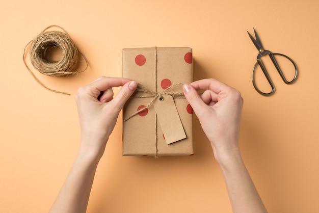 Erste-Person-Draufsichtfoto von weiblichen Händen im Valentinstag-Dekor, die eine Schleife auf einer Geschenkbox aus Kraftpapier mit Tupfenmuster-Preisschild binden