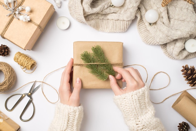 Erste-Person-Draufsichtfoto von weiblichen Händen in Pullovern, die Kraftpapier-Geschenkboxen mit Tannenzweigen, gestrickten Pullover-Weihnachtsbaumkugeln und Werkzeugen für das Handwerk auf isoliertem weißem Hintergrund dekorieren