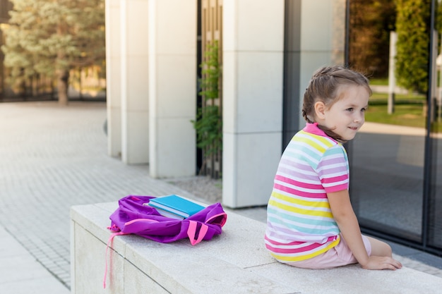 Erster Schultag. Schüler der Grundschule mit Buch in der Hand. Mädchen mit einem Rucksack nahe dem Gebäude im Freien.