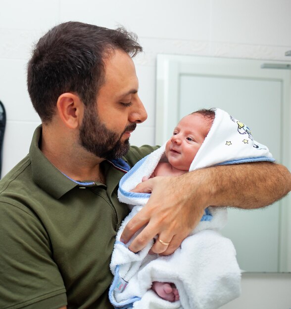 Erstes Bad des neugeborenen Jungen Das Baby liegt im Bademantel in den Armen seines Vaters