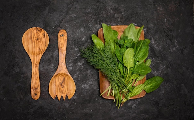 Ervas frescas espinafre azeda endro na placa de madeira com garfo de salada de madeira e colher em fundo preto Dieta vegana e conceito de alimentação limpa