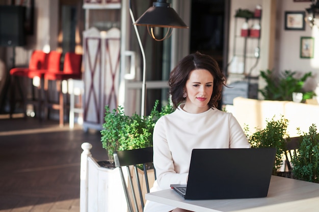 Erwachsene elegante Geschäftsfrau im weißen Kleid, das mit Laptop im Café sitzt. Schöne brünette Dame mit Notizbuch im Café. Schuss einer Frau mittleren Alters, die auf der Tastatur tippt