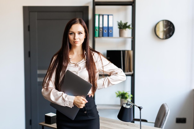 Erwachsene erfolgreiche weibliche Angestellte, die in einem modernen Büro stehen, halten einen Laptop in den Armen. Glückliche Geschäftsfrau mit brünettem Haar in elegantem, stilvollem Anzug blickt in die Kamera und lächelt