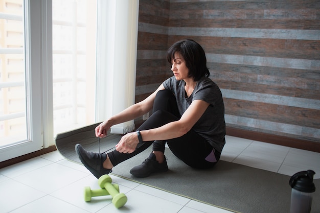 Erwachsene fit schlanke Frau hat Training zu Hause. Ernsthaft fleißiges reifes Modell sitzen auf Yogamatte, um Schnürsenkel zu binden. Vorbereitungen für das Training.