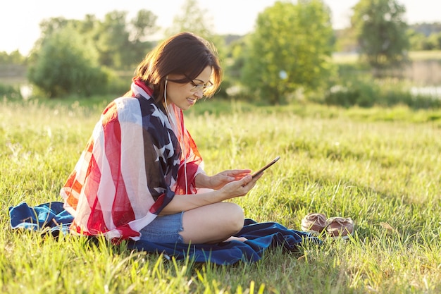 Erwachsene Frau, die am Juliabend in der Natur mit USA-Flaggenschal sitzt, Frau in Kopfhörern mit Smartphone, die Feier zum Unabhängigkeitstag sieht