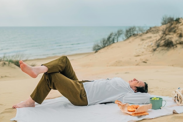 Erwachsene Frau im Frühjahr bei einem Picknick am Strand am Meer liegt auf Plaid mit einem Lächeln