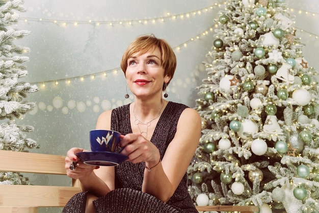 Erwachsene Frau im neuen Jahr auf der Eisbahn trinkt Kaffee auf einer Bank neben dem Weihnachtsbaum