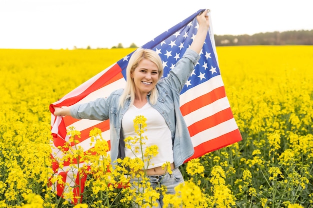 Erwachsene Frau mit amerikanischer Flagge mit Stange, Stars and Stripe in einem gelben Rapsfeld. USA-Flagge flattert im Wind.