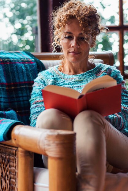 Erwachsene Frau mittleren Alters, die zu Hause ein Buch liest und bequem auf einem Liegestuhl sitzt. Indoor-Freizeitaktivität mit alleinstehenden weiblichen Menschen, die lächeln und sich entspannen