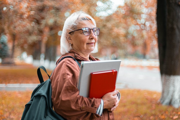 Erwachsene kaukasische blonde Lehrerfrau in Freizeitkleidung steht mit Notizbüchern und Rucksack, die im Herbstpark gehen