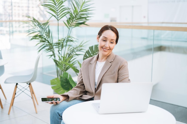 Erwachsene lächelnde brünette Geschäftsfrau vierzig Jahre mit langen Haaren in stylischem beigefarbenem Anzug und Jeans, die an einem Laptop im öffentlichen Büro arbeitet