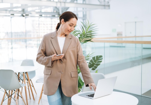 Erwachsene lächelnde brünette Geschäftsfrau vierzig Jahre mit langen Haaren in stylischem beigefarbenem Anzug und Jeans, die an einem Laptop im öffentlichen Büro arbeitet