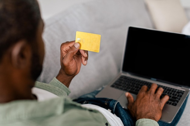 Erwachsener Afroamerikaner tippt lässig auf Laptop mit leerem Bildschirm und zeigt Kreditkarte