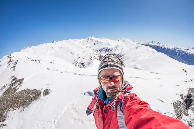 Erwachsener Alpin-Skifahrer mit Bart, Sonnenbrille und Hut, selfie auf schneebedeckter Steigung in den schönen italienischen Alpen mit klarem blauem Himmel nehmend. Konzept von Fernweh und Abenteuer am Berg