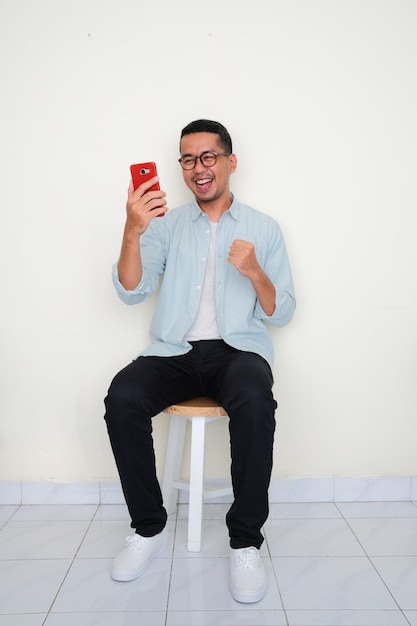 Erwachsener asiatischer Mann, der auf einer Bank sitzt und mit aufgeregtem Ausdruck auf sein Telefon schaut