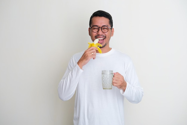 Erwachsener asiatischer Mann, der glücklichen Ausdruck zeigt, wenn er ein Glas Wasser hält und Banane isst