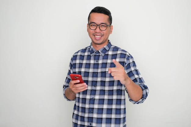 Erwachsener asiatischer Mann lächelt und zeigt auf die Kamera, während er ein Handy hält