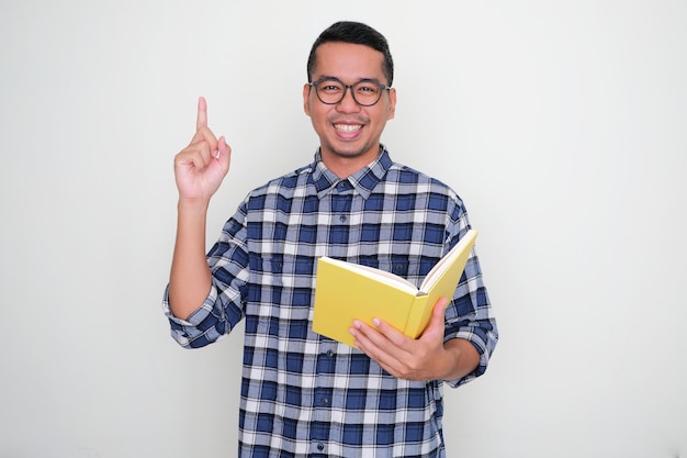 Erwachsener asiatischer Mann lächelt und zeigt mit dem Finger nach oben, während er ein Buch hält