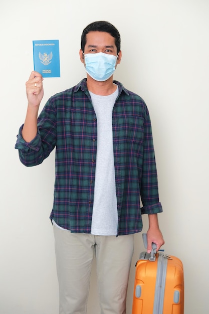 Erwachsener asiatischer Mann mit medizinischer Schutzmaske, der einen indonesischen Pass hält und Reisekoffer mitbringt