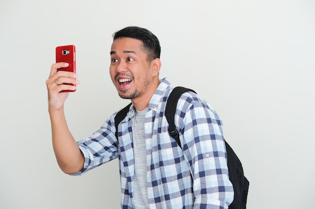 Erwachsener asiatischer Mann mit Rucksack, der aufgeregten Ausdruck zeigt, wenn er ein Foto mit dem Handy macht