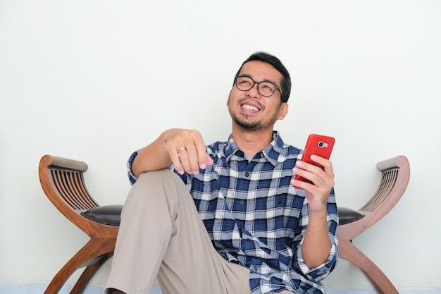 Erwachsener asiatischer Mann sitzt entspannt und hält Handtelefon, während er sich etwas vorstellt