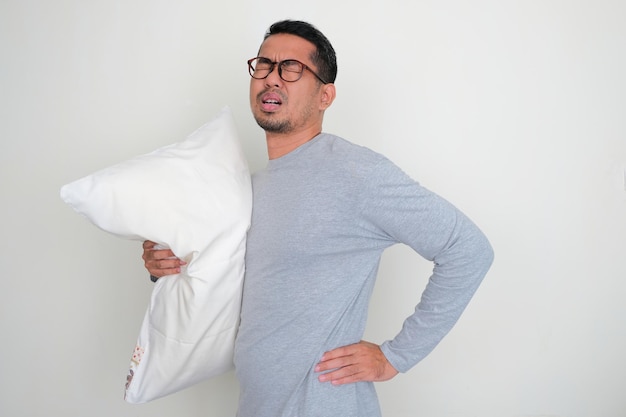 Erwachsener asiatischer Mann umarmt Schlafkissen, während er seinen Rücken mit schmerzlichem Ausdruck berührt