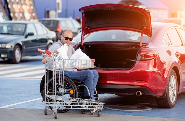 Erwachsener behinderter Mann im Rollstuhl legt Einkäufe in den Kofferraum eines Autos auf einem Supermarktparkplatz