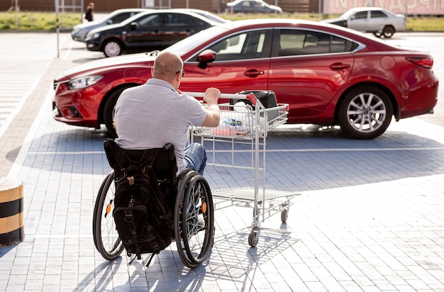 Erwachsener behinderter Mann im Rollstuhl schiebt einen Wagen auf einem Supermarktparkplatz auf ein Auto zu