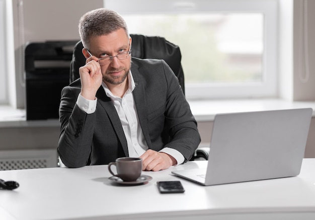 Erwachsener erfolgreicher Mann in einem grauen Business-Anzug, der an einem Tisch sitzt und eine Brille hält