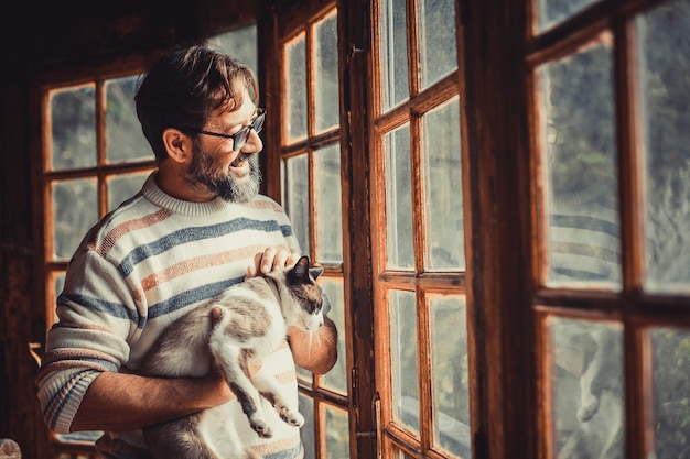 Erwachsener glücklicher reifer Mann mit einer Katze schaut zu Hause vor den Fenstern und genießt allein die Entspannung und Aktivität im Innenbereich. Hipster-Männer mit Fensterlicht, die im Wohnzimmer lächeln