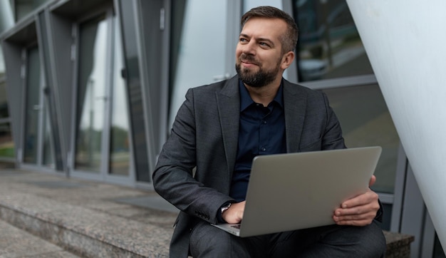 Erwachsener gutaussehender Geschäftsmann, der online am Laptop neben der Fassade des Büro-Business-Center-Konzepts arbeitet