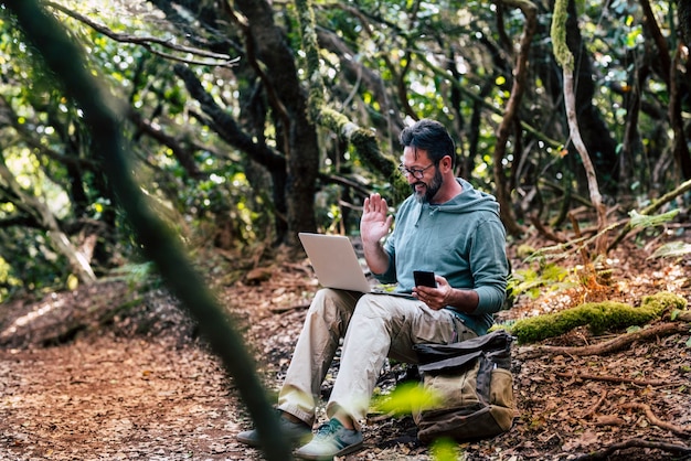 Erwachsener kaukasischer Mann, der auf dem Boden im Wald sitzt, führt eine Videoanrufverbindung mit einem Laptop-Computer durch - drahtloses Internet überall und Telefon-Roaming-Hot-Spot-Konzept