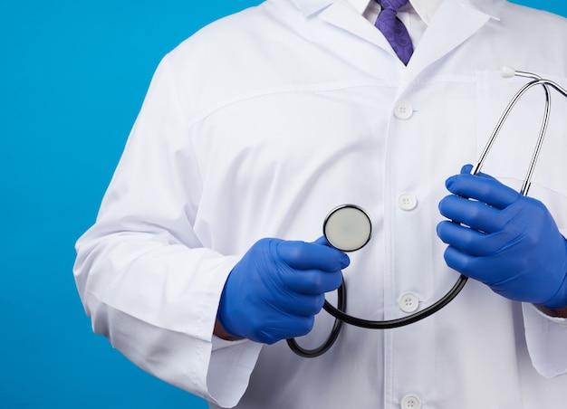 Erwachsener männlicher Arzt in einem weißen medizinischen Mantel steht und hält ein schwarzes Stethoskop auf einem blauen Hintergrund
