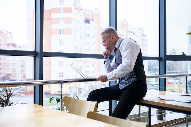 Erwachsener männlicher Geschäftsmann, Lehrer, Mentor, der an einem neuen Projekt arbeitet. Sitzt an einem großen Fenster auf dem Tisch. Er schaut auf den Bildschirm des Laptops.