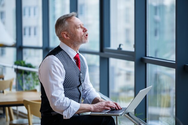 Erwachsener männlicher Geschäftsmann, Lehrer, Mentor, der an einem neuen Projekt arbeitet. Sitzt an einem großen Fenster auf dem Tisch. Er schaut auf den Bildschirm des Laptops.