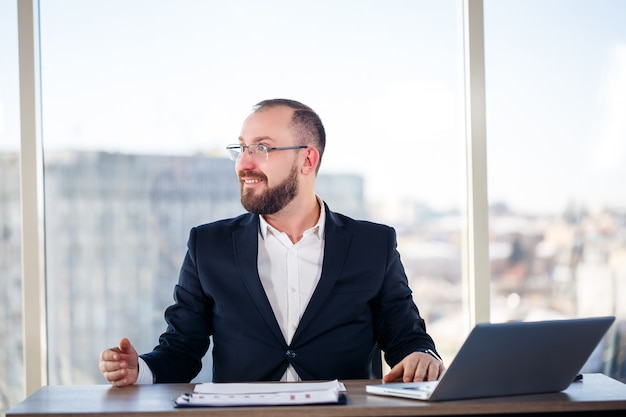 Erwachsener männlicher Mentor, Direktor, Geschäftsmann in Brille und Anzug, der Dokumente studiert, während er am Tisch sitzt. Konzept des Arbeitstages