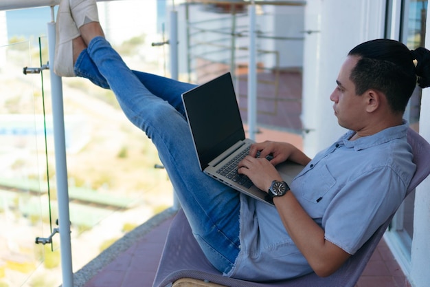 Erwachsener Mann, der am Laptop arbeitet, während er auf dem Balkon seines Hauses sitzt