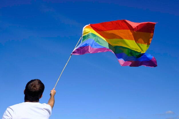 Erwachsener Mann im weißen T-Shirt winkt Regenbogenfahne mit blauem Himmel.